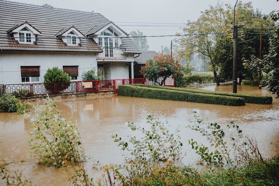 A flooded residential street during a severe weather event, with water covering the pavement and partially submerging parked vehicles, including a silver car and several others in the background. Surr