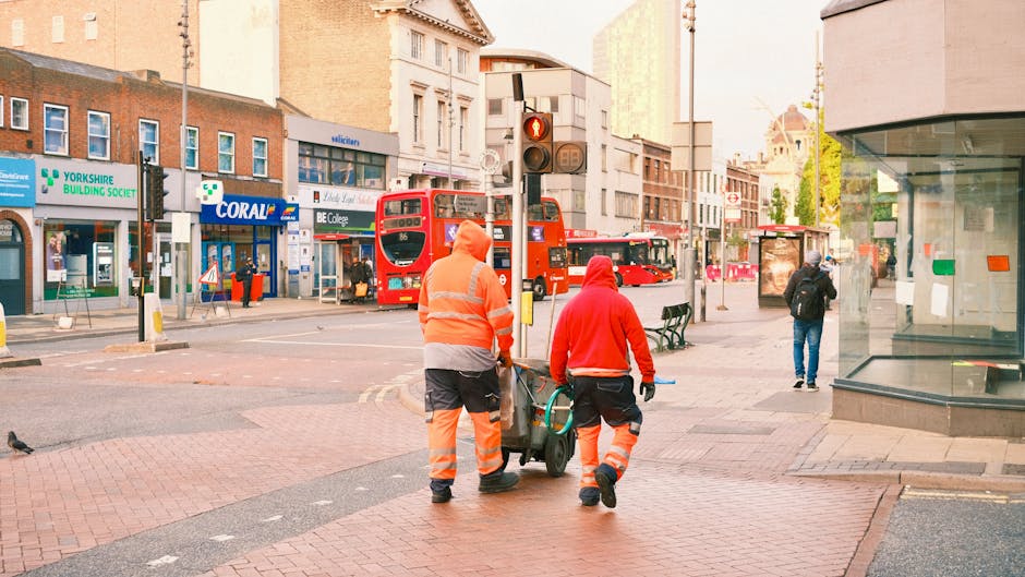 Two street cleaning workers wearing orange and red high-visibility jackets and trousers are seen walking along a pedestrian crossing on Hounslow High Street, near a glass-fronted shop. They are pushing a cleaning trolley equipped with brushes and cleaning tools. In the background, there are red double-decker buses, various storefronts including a Yorkshire Building Society branch and a shop named Coral, and a traffic light showing red. Some pedestrians are walking further down the street, and sunlight illuminates the scene, highlighting the cleanliness and orderliness of the urban area. The scene emphasizes routine surface cleaning and maintenance, reflecting the services offered by Cleaners Hounslow for domestic and commercial cleaning needs.