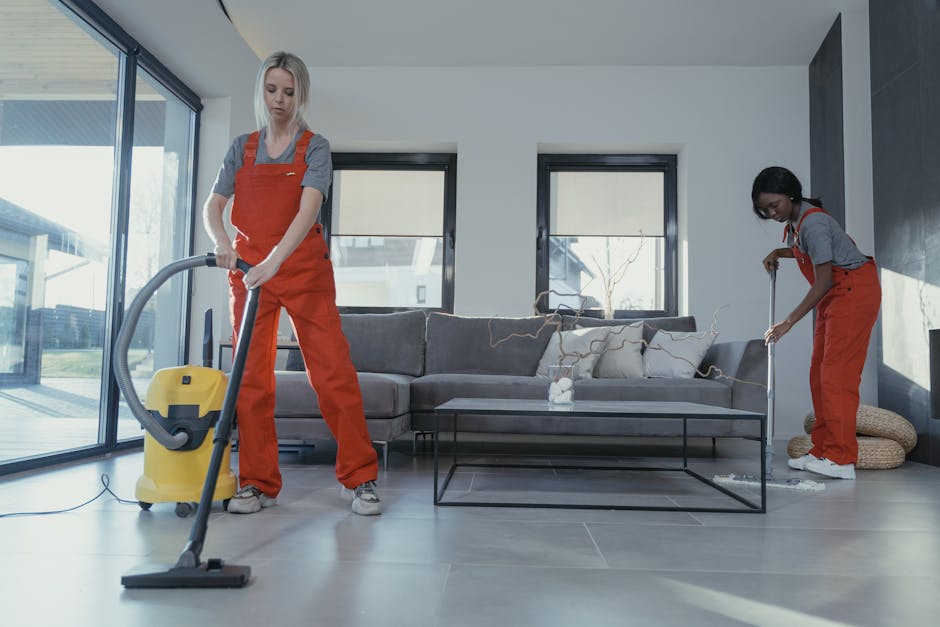 A professional cleaner from Cleaners Hounslow is in a modern kitchen, operating a yellow industrial vacuum cleaner for surface cleaning and deep cleaning. The cleaner, a woman with dark hair, is dress