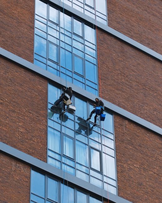 Two professional window cleaners from Cleaners Hounslow are performing high-rise surface cleaning on the exterior glass façade of a modern brick building. They are seated on a suspended platform, equ